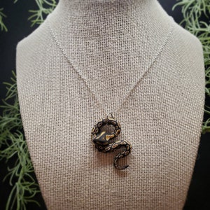 Op de afbeelding: Een zilveren ketting met een hanger in de vorm van een opgerolde slang. De slangenhanger is zwart met bruine en gouden markeringen. De ketting wordt getoond op een beige sieraden buste tegen een donkere achtergrond met groen gebladerte.