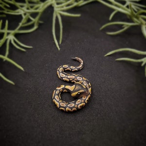 Op de afbeelding: Een gedetailleerde miniatuursculptuur van een opgerolde slang. De slang heeft een patroon van zwarte, bruine en gele markeringen. De sculptuur staat tegen een zwarte achtergrond, met groen gebladerte op de achtergrond.
