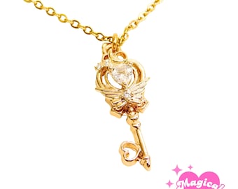 May include: A gold-toned necklace featuring a key-shaped pendant with a heart-shaped center, small clear stones, and wing-like accents. The bottom of the key has a heart-shaped bow. A pink heart logo with the text Magical Girl Me is visible.