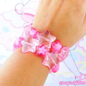 Può includere: Un braccialetto rosa a forma di stella con perline rosa trasparenti e piene. Il braccialetto è indossato su un polso, mostrando il design e il colore dell'accessorio. Un accessorio divertente e giocoso.