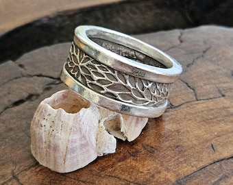 Puede incluir: Un anillo de plata con una banda ancha que presenta un diseño floral detallado. El anillo está apoyado en una concha.