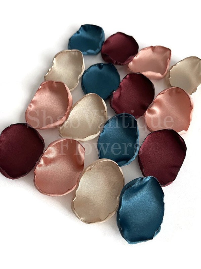 May include: Assortment of silk flower petals in burgundy, blush pink, cream, and teal colors. The petals are arranged on a white surface.