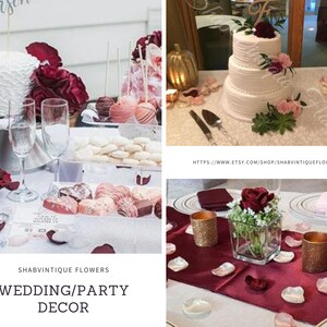 May include: A wedding or party decor table setting with a white cake decorated with burgundy and pink flowers, a bouquet of burgundy and pink roses, and a centerpiece with a single burgundy rose in a glass vase. The table is decorated with burgundy and pink rose petals.