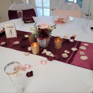 Puede incluir: Una mesa redonda blanca preparada para un evento formal con un camino de mesa de sat&eacute;n burdeos, un centro de mesa floral en un jarr&oacute;n de purpurina plateada, dos portavelas de purpurina dorada, p&eacute;talos de rosa y una tarjeta de mesa n&uacute;mero 8.