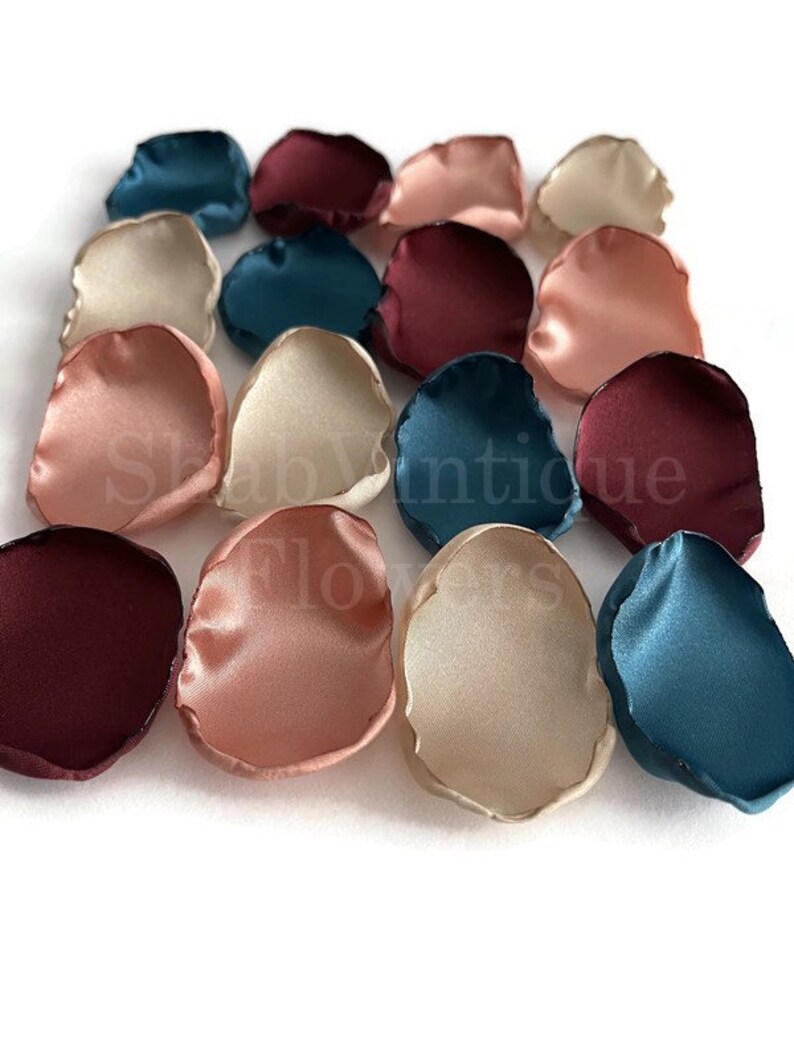 May include: A collection of silk flower petals in shades of burgundy, teal, and blush. The petals are arranged in a random pattern on a white background.