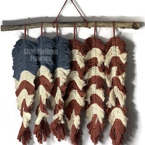 Puede incluir: Un tapiz de pared hecho de macramé con colores rojo, blanco y azul. El macramé está colgado de una rama y tiene una franja en la parte inferior.