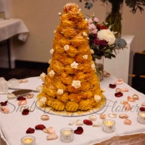 May include: A tall, cone-shaped wedding cake made of cream puffs, decorated with white flowers and a white ribbon. The cake is on a white table with red and pink rose petals scattered around it.