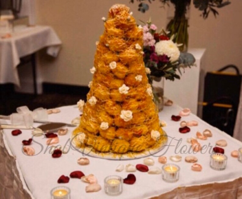 May include: A tall, cone-shaped wedding cake made of cream puffs, decorated with white flowers and a white ribbon. The cake is on a white table with red and pink rose petals scattered around it.