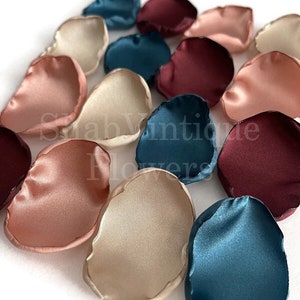 May include: Assortment of silk rose petals in blush pink, burgundy, teal, and ivory colors. The petals are scattered on a white surface.
