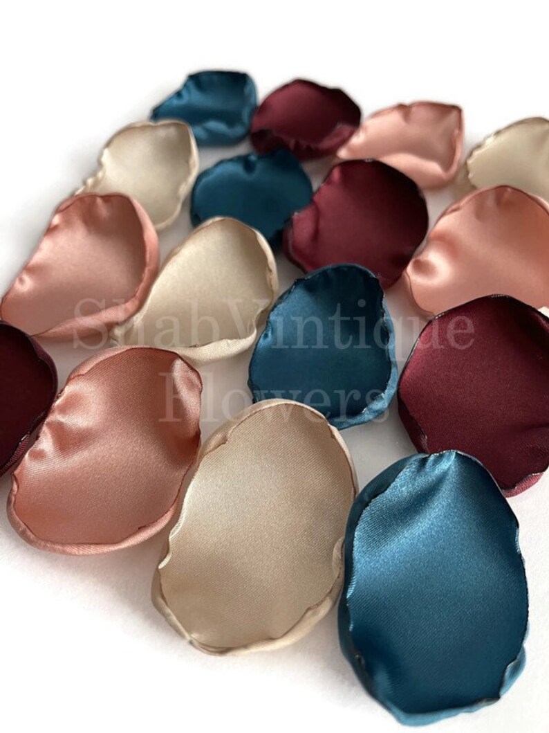 May include: Assortment of silk rose petals in blush pink, burgundy, teal, and ivory colors. The petals are scattered on a white surface.