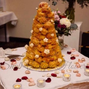 Puede incluir: Un postre croquembouche c&oacute;nico construido con profiteroles dorados unidos con az&uacute;car hilado, decorado con peque&ntilde;os detalles florales blancos y colocado sobre un mantel blanco esparcido con p&eacute;talos de rosa rojos y rosas y velas de t&eacute;.