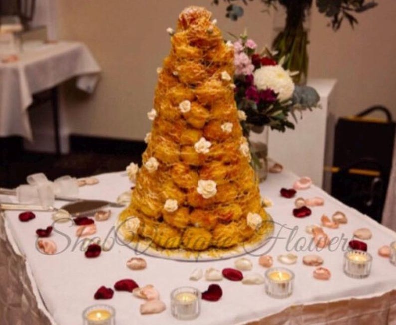 Puede incluir: Un postre croquembouche c&oacute;nico construido con profiteroles dorados unidos con az&uacute;car hilado, decorado con peque&ntilde;os detalles florales blancos y colocado sobre un mantel blanco esparcido con p&eacute;talos de rosa rojos y rosas y velas de t&eacute;.