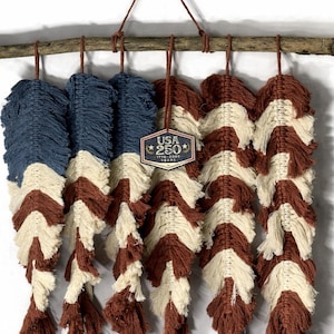 May include: A macrame wall hanging with feather-shaped designs in navy, cream, and brown, suspended from a wooden branch. The piece includes a tag with the text "USA 250". The design uses colours reminiscent of a flag.