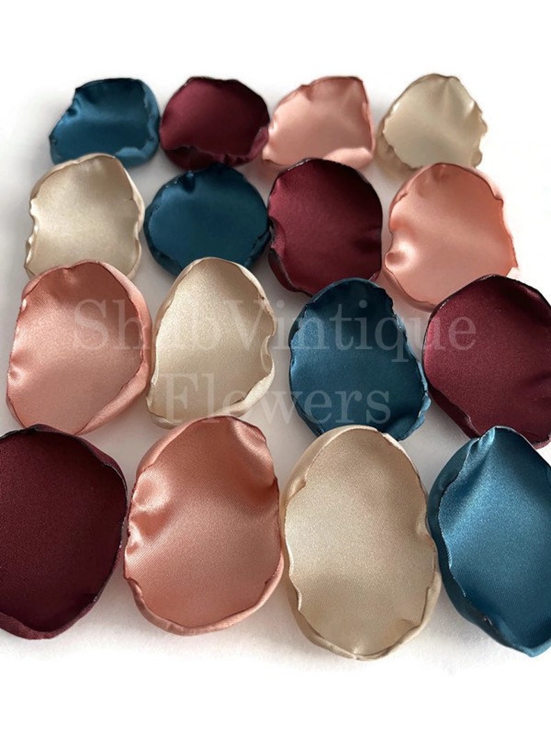 May include: Assortment of silk flower petals in burgundy, teal, blush, and ivory colors. The petals are arranged in a random pattern on a white surface.