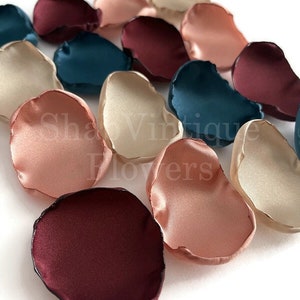 May include: Assortment of silk flower petals in burgundy, blush pink, ivory, and teal colors. The petals are arranged in a row on a white surface.