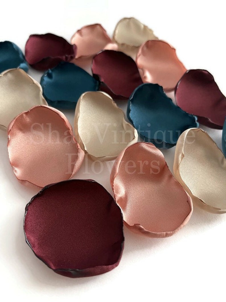 May include: Assortment of silk flower petals in burgundy, blush pink, ivory, and teal colors. The petals are arranged in a row on a white surface.