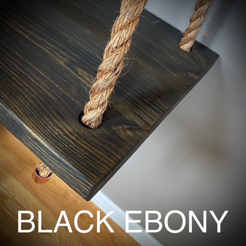 Puede incluir: Un estante rectangular de madera te&ntilde;ido en un acabado negro &eacute;bano con veta de madera visible. El estante est&aacute; suspendido por una cuerda de yute trenzada de color natural que pasa a trav&eacute;s de orificios circulares. Un accesorio de metal cobrizo.