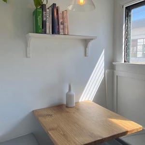May include: A wall-mounted wooden shelf holding a row of books, positioned above a square wooden table. A white ceramic vase sits on the table surface. A glass pendant light hangs from the ceiling, and a window is visible on the right side.