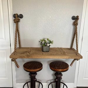 May include: A wall-mounted floating shelf suspended by thick natural fiber ropes attached to dark metal pipe fixtures. The rectangular wooden shelf holds a small metal pot with artificial flowers on a woven mat, paired with two round wooden stools.