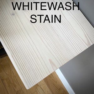 Puede incluir: Una tabla de madera rectangular con un acabado de tinte blanco que resalta la veta y la textura natural de la madera. El texto WHITEWASH STAIN est&aacute; impreso en letras may&uacute;sculas negras en la parte superior de la superficie.