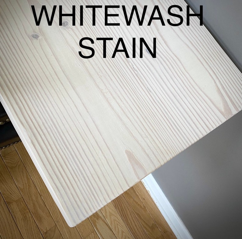 Puede incluir: Una tabla de madera rectangular con un acabado de tinte blanco que resalta la veta y la textura natural de la madera. El texto WHITEWASH STAIN est&aacute; impreso en letras may&uacute;sculas negras en la parte superior de la superficie.