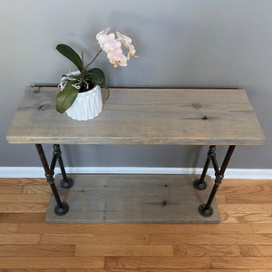 May include: A gray wooden console table with a black metal pipe frame. The table has a single shelf and is decorated with a potted orchid.