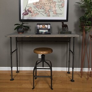 May include: A gray wooden desk with black metal pipe legs and a black metal adjustable stool. The desk has a map of Hong Kong hanging above it.