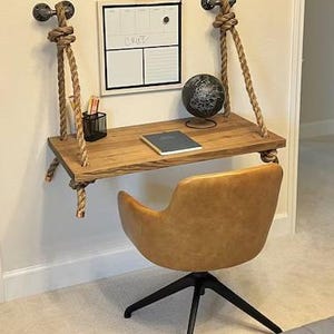Puede incluir: Un escritorio flotante montado en la pared hecho de una tabla de madera rectangular suspendida por gruesas cuerdas color canela unidas a accesorios de tuber&iacute;a de metal negro. Una silla giratoria tapizada en color canela con base negra se sit&uacute;a frente al escritorio, que sostiene un globo terr&aacute;queo, un cuaderno y un portal&aacute;pices de alambre negro. Una pizarra blanca con el texto "CRUZ" est&aacute; montada encima.