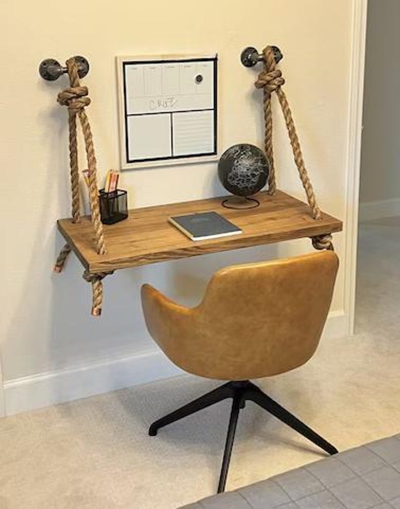 Puede incluir: Un escritorio flotante montado en la pared hecho de una tabla de madera rectangular suspendida por gruesas cuerdas color canela unidas a accesorios de tuber&iacute;a de metal negro. Una silla giratoria tapizada en color canela con base negra se sit&uacute;a frente al escritorio, que sostiene un globo terr&aacute;queo, un cuaderno y un portal&aacute;pices de alambre negro. Una pizarra blanca con el texto "CRUZ" est&aacute; montada encima.