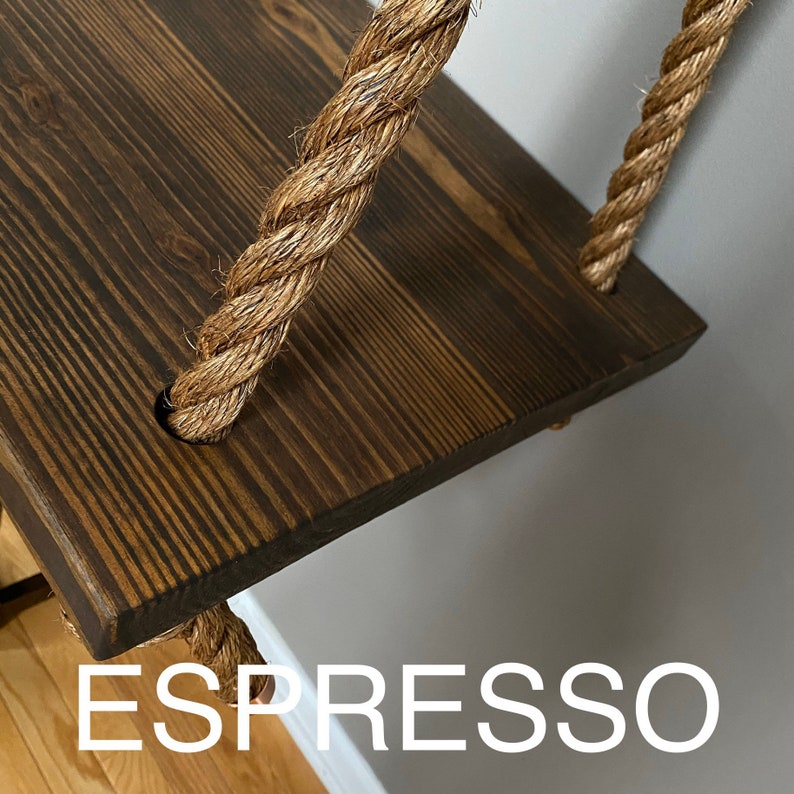 Puede incluir: Un estante de madera te&ntilde;ido de marr&oacute;n oscuro suspendido por gruesas cuerdas de fibra natural trenzada. El estante rectangular presenta una veta de madera visible y est&aacute; asegurado por cuerdas que pasan por orificios circulares. La palabra ESPRESSO aparece debajo.