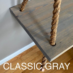 Puede incluir: Un estante rectangular de madera te&ntilde;ido en un acabado gris cl&aacute;sico, suspendido por gruesas cuerdas de yute trenzado de color natural que pasan a trav&eacute;s de orificios circulares en cada esquina. La superficie muestra la veta de la madera.