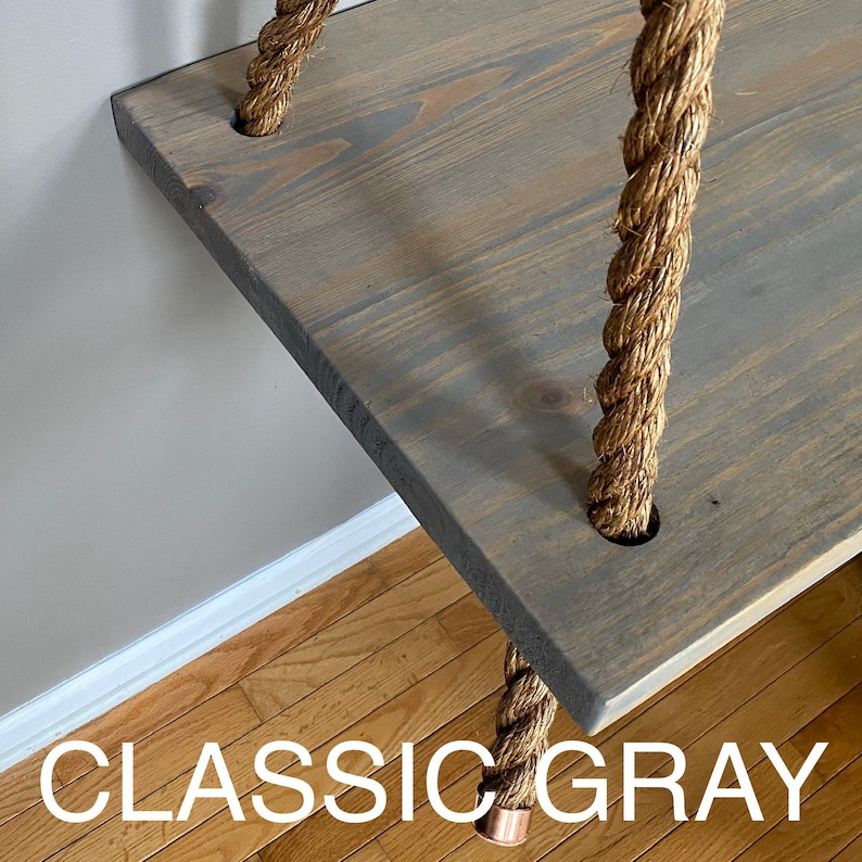 Puede incluir: Un estante rectangular de madera te&ntilde;ido en un acabado gris cl&aacute;sico, suspendido por gruesas cuerdas de yute trenzado de color natural que pasan a trav&eacute;s de orificios circulares en cada esquina. La superficie muestra la veta de la madera.