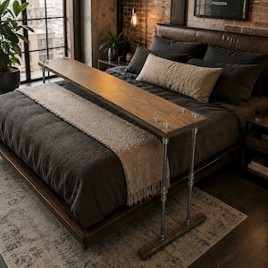 May include: A wooden bed tray with metal legs extends over a bed with dark gray bedding. The tray is made of light-colored wood and is attached to the metal legs with silver bolts. The bed is in a room with a brick wall and a large window.
