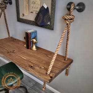 Puede incluir: Un estante rectangular de madera con acabado de veta marr&oacute;n oscuro, suspendido por gruesas cuerdas de fibra natural. Las cuerdas est&aacute;n aseguradas a la pared con accesorios de tuber&iacute;a de metal industrial gris oscuro y tapas de cobre.