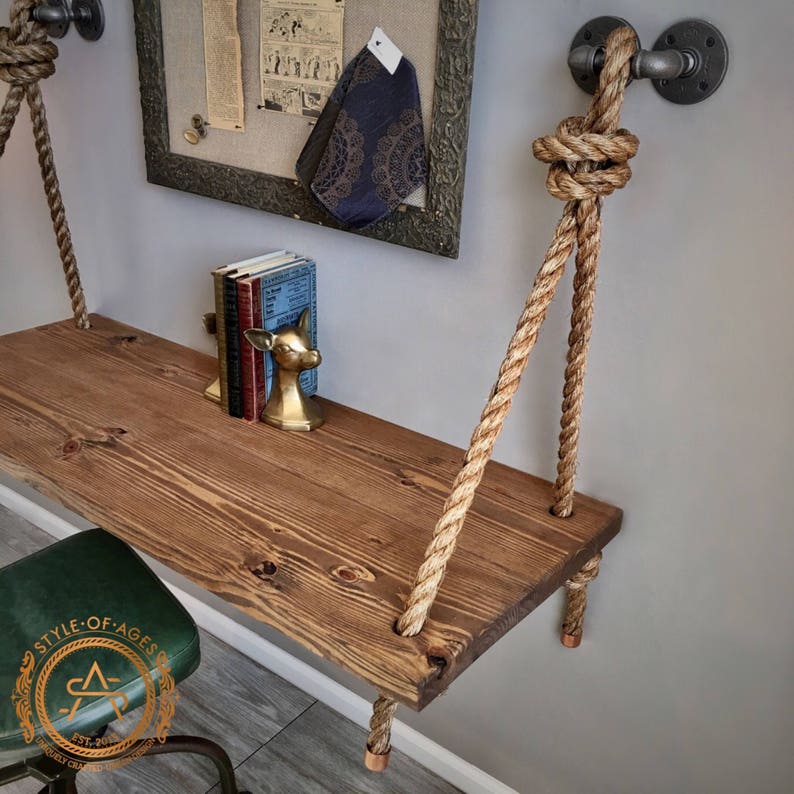 Puede incluir: Un estante rectangular de madera con acabado de veta marr&oacute;n oscuro, suspendido por gruesas cuerdas de fibra natural. Las cuerdas est&aacute;n aseguradas a la pared con accesorios de tuber&iacute;a de metal industrial gris oscuro y tapas de cobre.