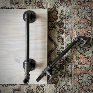 May include: Two black metal industrial style door handles with pipe fittings. The handles are mounted on a white wooden surface.