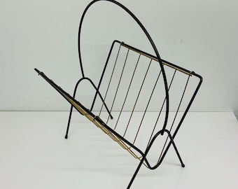 May include: A mid-century modern magazine rack. The rack is made of black metal with gold-colored accents. It has a curved handle and a series of vertical bars to hold magazines. The rack stands on four thin legs.