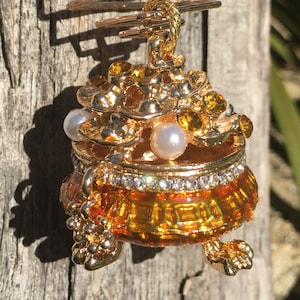 Puede incluir: Un adorno dorado con un recipiente de vidrio transparente decorado con diamantes de imitación dorados y transparentes. El recipiente está unido a una espiral dorada y tiene una perla en su interior.