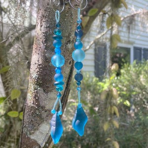 Puede incluir: Un par de pendientes hechos a mano con un diseño en cascada. Los pendientes presentan una mezcla de cuentas de vidrio azul y colgantes en forma de lágrima. Los ganchos para las orejas son de color plateado.