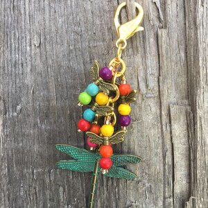 Puede incluir: Un llavero de libélula colorido con un cierre dorado y cuentas en tonos turquesa, amarillo, rojo, morado y verde.
