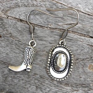 May include: Silver-toned cowboy boot and hat earrings. The earrings feature a detailed cowboy boot charm and a cowboy hat charm with a rope-like border. The earrings are displayed on a weathered wooden surface.