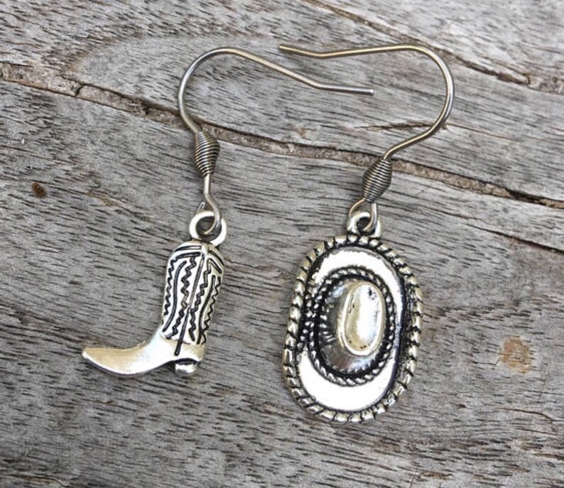 May include: Silver-toned cowboy boot and hat earrings. The earrings feature a detailed cowboy boot charm and a cowboy hat charm with a rope-like border. The earrings are displayed on a weathered wooden surface.