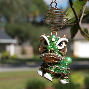 May include: A detailed, green and gold lion dance ornament with white accents and a spiral hanging wire. The ornament features a detailed design with green and gold enamel, rhinestones, and pearl accents. The ornament is hanging from a tree branch.