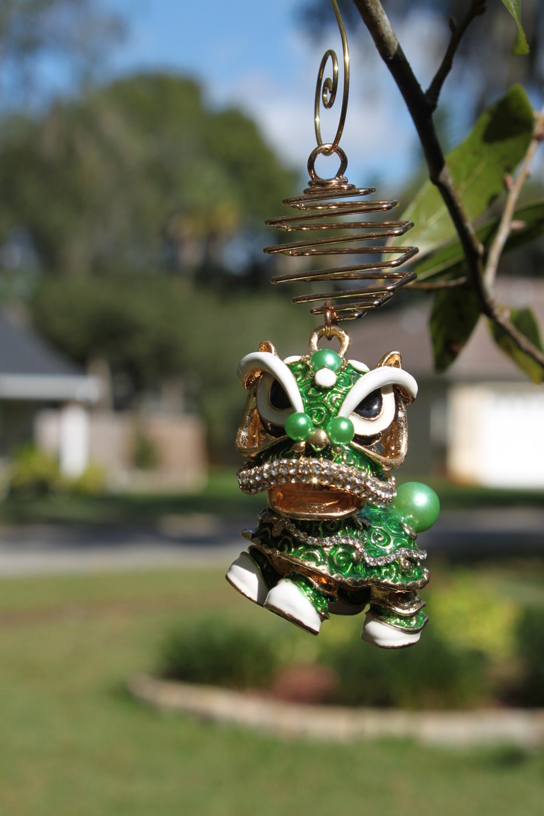May include: A detailed, green and gold lion dance ornament with white accents and a spiral hanging wire. The ornament features a detailed design with green and gold enamel, rhinestones, and pearl accents. The ornament is hanging from a tree branch.