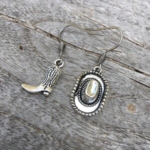 May include: Silver-toned cowboy boot and hat earrings. The boot charm has detailed stitching, and the hat charm features a rope-like border. The earrings hang from silver-colored fish hook ear wires. The earrings are displayed on a weathered wooden surface.