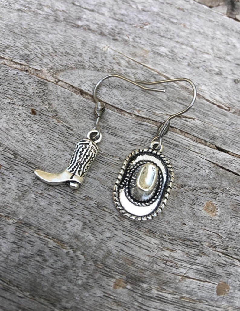 May include: Silver-toned cowboy boot and hat earrings. The boot charm has detailed stitching, and the hat charm features a rope-like border. The earrings hang from silver-colored fish hook ear wires. The earrings are displayed on a weathered wooden surface.