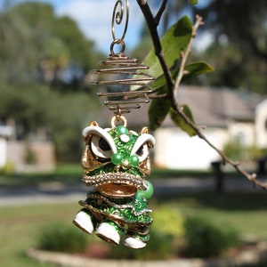 May include: A decorative ornament featuring a green and gold lion dance design, suspended from a gold wire. The ornament has white accents, faux gems, and is hanging from a tree branch against a blurred outdoor background.