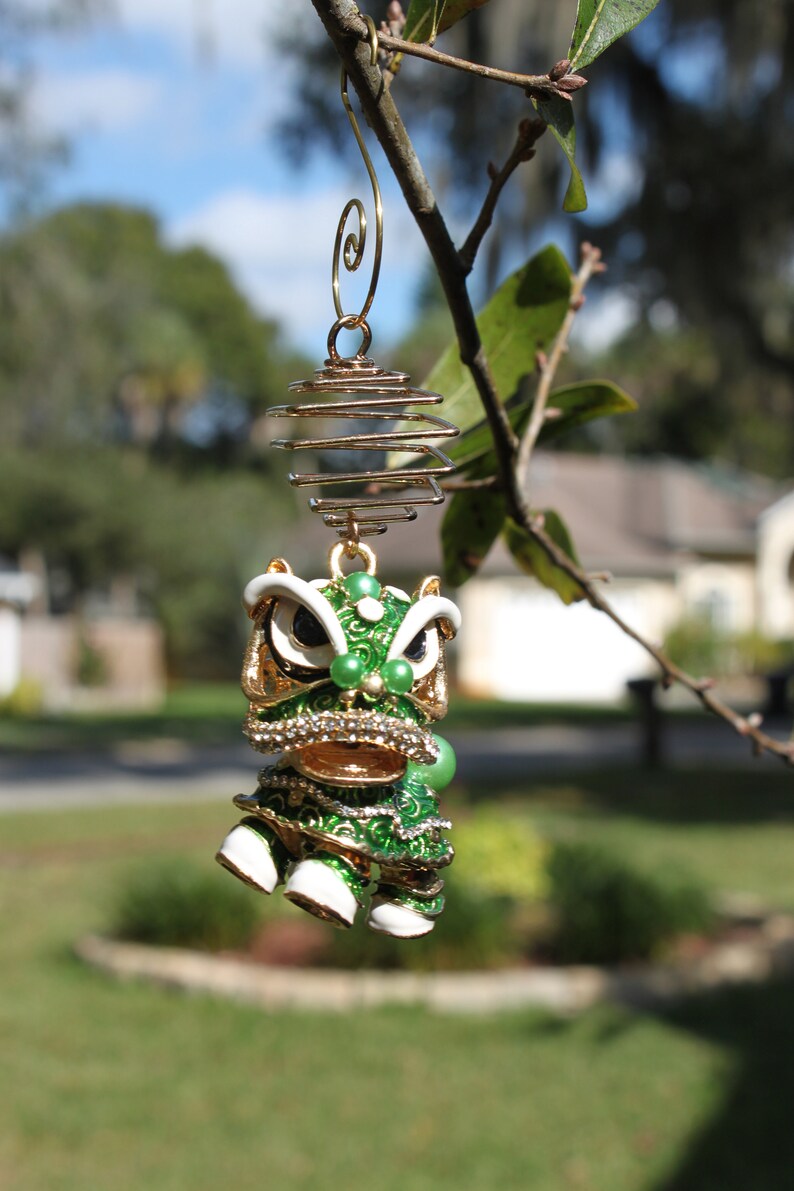 May include: A decorative ornament featuring a green and gold lion dance design, suspended from a gold wire. The ornament has white accents, faux gems, and is hanging from a tree branch against a blurred outdoor background.