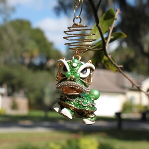 May include: A detailed, green and gold lion dance ornament with white accents and sparkling rhinestones. The ornament is suspended from a gold wire spiral and a tree branch, set against a blurred outdoor background.