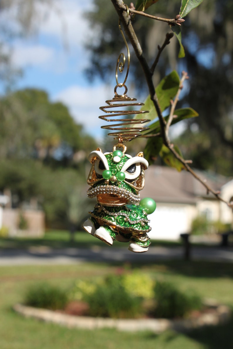 May include: A detailed, green and gold lion dance ornament with white accents and sparkling rhinestones. The ornament is suspended from a gold wire spiral and a tree branch, set against a blurred outdoor background.